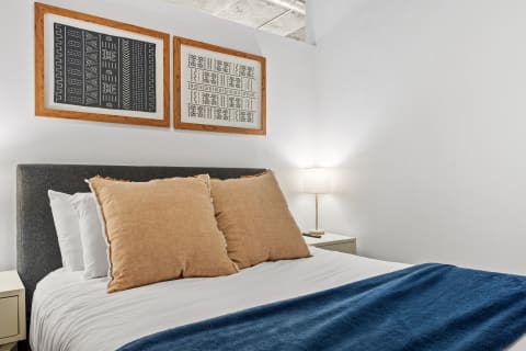 Cozy bedroom with a gray headboard, large beige pillows, blue throw blanket, and art on the wall.