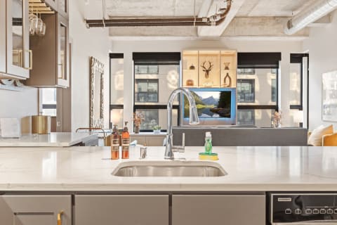 A stylish kitchen with marble countertop, chrome faucet, and a view of a living room with a TV showing a landscape.