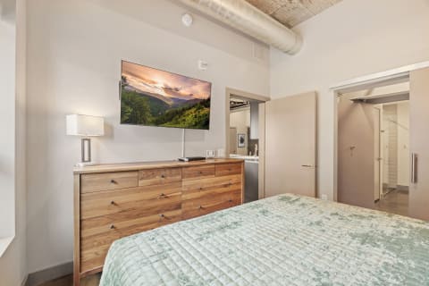A modern bedroom with a green bedspread, wooden dresser, and a flat-screen TV showing a landscape.