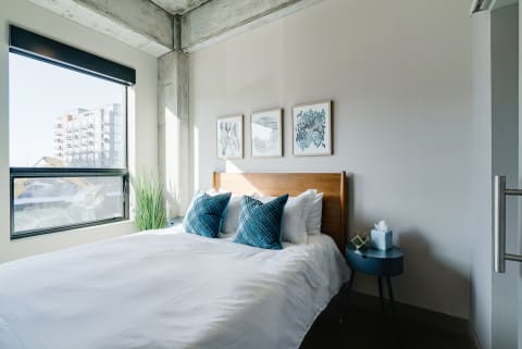 A stylish bedroom with white bedding, turquoise pillows, abstract art, and a window overlooking an urban landscape.