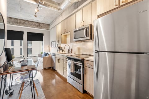 Modern studio apartment with kitchen and living area featuring light wood cabinetry and stainless steel appliances.
