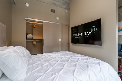 A cozy modern bedroom with a white comforter, sliding closet, and a television displaying the MINNESTAY logo.