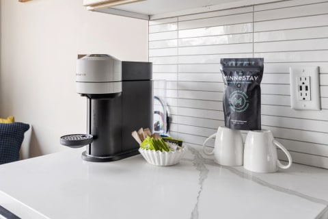 A countertop with a Nespresso coffee machine, white mugs, and coffee condiments in a bowl, set against a tiled background.