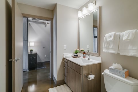 Modern bathroom with a wooden vanity, mirror, and towel rack, leading to another room.
