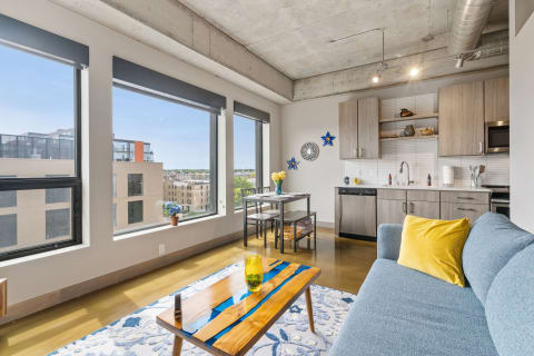 Interior view of a modern apartment featuring a cozy blue sofa, wooden table, and large windows with city views.
