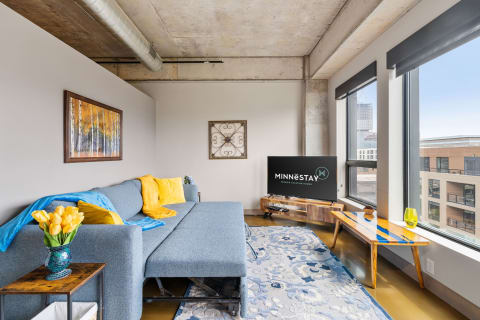 Contemporary living room with blue couch and yellow pillows, featuring a large window and TV.