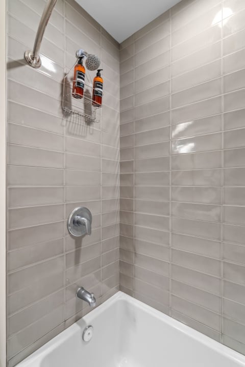 A contemporary shower area featuring light gray tiles, a showerhead, and bottles of shampoo and conditioner.
