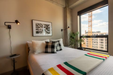 Cozy bedroom featuring a bed with white bedding and checkered pillows, a striped blanket, and a large window with a construction view.
