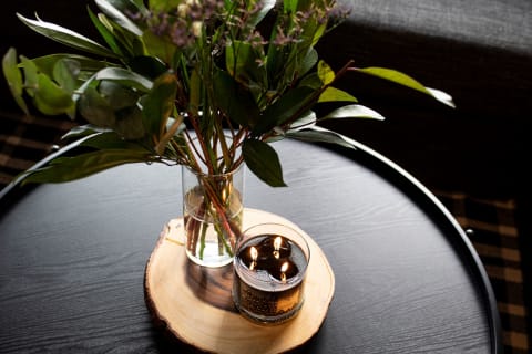 A glass vase with greenery and a lit candle on a wooden slice on a dark table.
