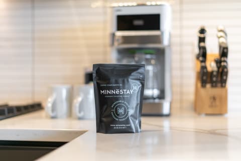 A black coffee bag for MINNESTAY displayed on a marble kitchen countertop with a coffee maker and knife block in the background.