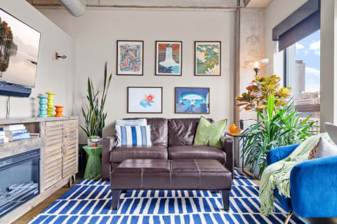 A modern living room featuring a brown leather sofa, colorful artwork, and lively plants.