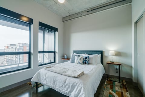 Modern bedroom with a bed, decorative pillows, bedside lamp, large windows, and an urban view.