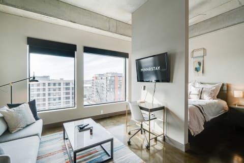 A modern studio apartment featuring a sofa, desk, bed, and large windows with city views.