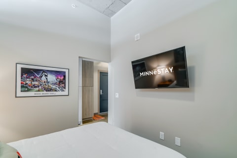 Contemporary guest room with a bed, TV displaying 'MINNESTAY,' and framed artwork.