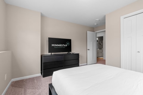 A cozy bedroom with a flat-screen TV on a black dresser, featuring a white bedspread and beige walls.