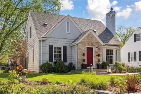 A two-story house featuring wooden siding, a stone entryway, and a red front door, surrounded by greenery.