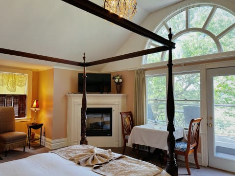 An elegant bedroom featuring a four-poster bed, a fireplace, and large windows overlooking a garden.