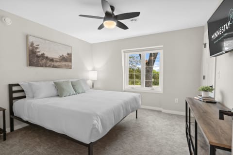 Modern bedroom featuring a bed with white linens, light green pillows, a ceiling fan, and a view of trees through a large window.