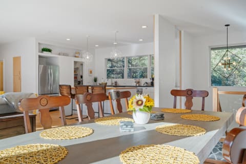 A bright dining room with a long wooden table, woven placemats, and a flower vase, transitioning into a kitchen area.