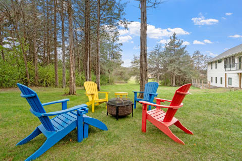 A backyard scene featuring colorful Adirondack chairs and a fire pit surrounded by trees.
