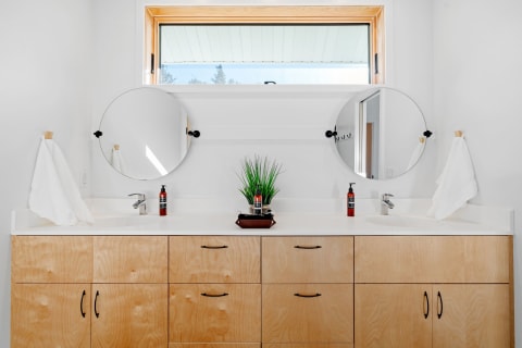Minimalistic bathroom with round mirrors, light wood cabinets, and a faux plant on the countertop.