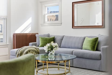 A living room with a grey sectional sofa, green pillows, a glass coffee table, and a vintage-style radiator.