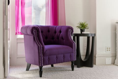 A plush purple armchair beside a sleek black side table with a potted plant, illuminated by natural light filtering through pink curtains.