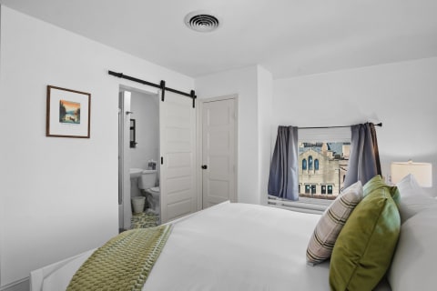 A cozy bedroom featuring a bed with green and patterned pillows, a sliding door to a bathroom, and a view of historic architecture through the window.