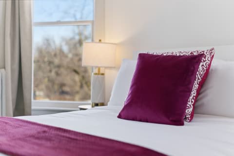 A stylish bedroom featuring a purple pillow on a white bed with a lamp nearby and soft curtains.