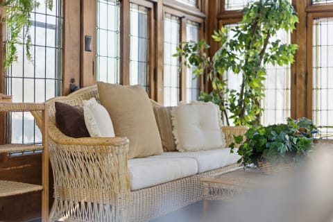 A wicker couch with cushions, surrounded by greenery and large windows.