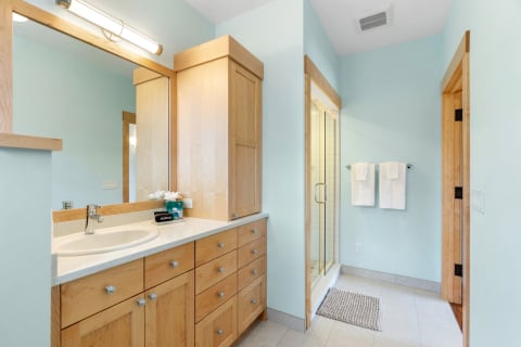 Modern bathroom with light blue walls, wooden vanity, and glass shower enclosure.