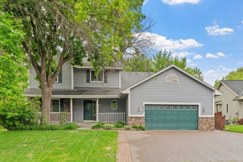 A two-story house with a front porch, green door, and brick accents, set in a leafy neighborhood.