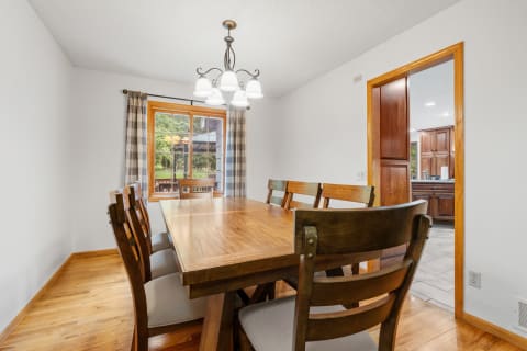 A dining room featuring a large wooden table, cushioned chairs, and a stylish chandelier with a view of greenery outside.