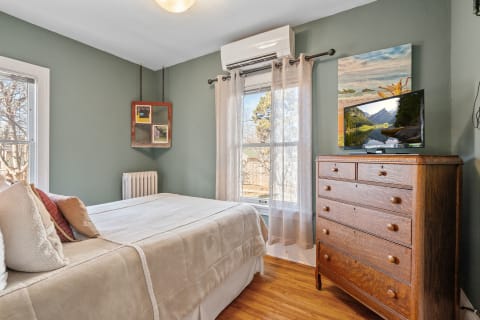 Cozy bedroom with green walls, a bed with beige covers, a wooden dresser, and artwork.