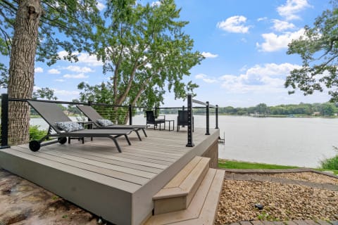 A modern wooden deck by the lake with two loungers and a table set, surrounded by trees and water.