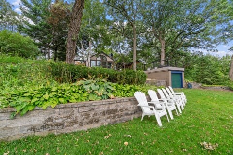 Outdoor scene featuring a row of white chairs, stone wall, and lush green garden with a house in the background.