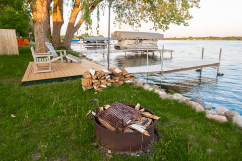 Lakeside view featuring a fire pit, wooden deck with chairs, and boats on a dock.