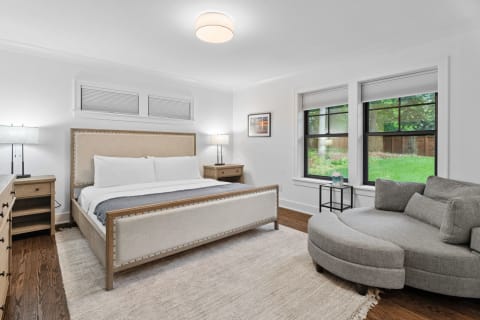 A modern bedroom with a bed, gray sofa, and view of a green backyard through large windows.