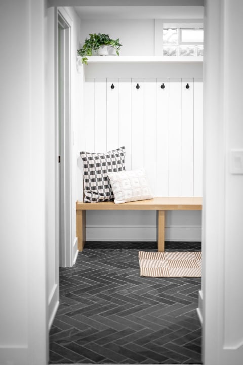 A minimalist entryway with a wooden bench, two pillows, and black herringbone flooring.