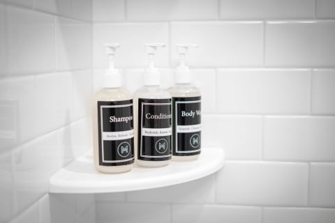 Three bottles labeled shampoo, conditioner, and body wash on a white shelf in a bathroom shower.