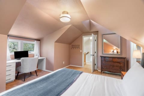 A cozy attic bedroom featuring sloped ceilings, a desk with a computer, a vintage dresser, and a neatly made bed.