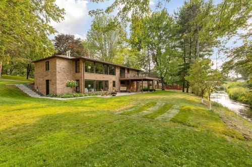 Modern brick home nestled among mature trees with expansive lawn and scenic river views beyond.