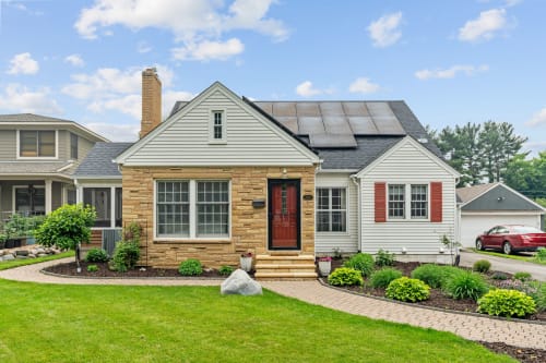 Charming stone and siding home with solar panels features a welcoming red front door and beautifully landscaped garden beds with mature trees.