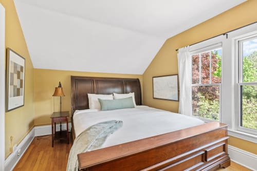 Rest peacefully in your serene bedroom with warm golden walls, elegant wooden furnishings, and natural light streaming through windows overlooking lush greenery.
